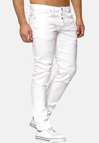 Jean en denim blanc avec une coupe slim, présentant des détails déchirés et plusieurs boutons sur le devant. Associé à des baskets blanches.