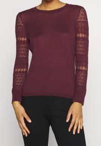 Burgunderfarbener Strickpullover mit langen Ärmeln; Spitzenakzente an den Ärmeln; gerippter Halsausschnitt und Bündchen; figurbetonte Silhouette; weiche Textur.