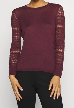 Pull en tricot bordeaux avec des manches longues ; détail en dentelle sur les manches ; encolure et poignets côtelés ; silhouette ajustée ; texture douce.