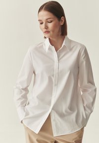 Chemise blanche en coton à manches longues avec un col classique et une fermeture boutonnée à l'avant. Présente une coupe décontractée et une texture lisse. Associée à un pantalon beige.