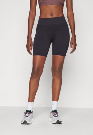 Frau trägt schwarze hoch taillierte Biker-Shorts, weiße Kniestrümpfe und weiße Sportschuhe mit rosa und grünen Akzenten, steht vor einem schlichten Hintergrund.