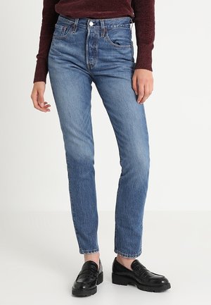 Frau trägt blaue High-Waist Skinny Jeans, schwarze Lederloafers und einen dunkelroten Langarmpullover, steht vor weißem Hintergrund.