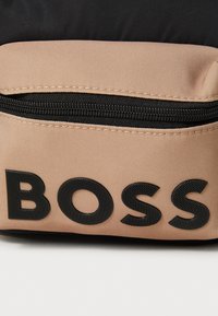 Črna in bež torba iz blaga, ki se ponaša z izstopajočim črnim logotipom "BOSS", z zadrgo na bež delu in teksturirano površino.