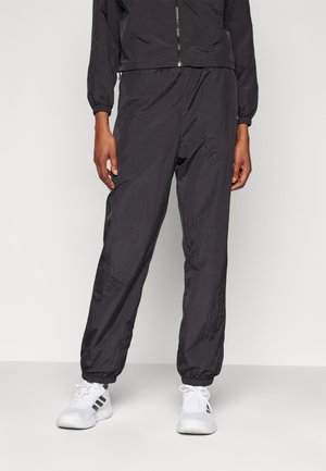 Persoon draagt zwarte elastische broek met taille en manchetten, gecombineerd met witte sportschoenen met zwarte strepen, staand tegen een witte achtergrond.