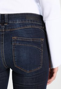 Jeans de denim azul oscuro con una silueta ajustada, que cuentan con bolsillos traseros y costuras naranjas en contraste, llevados con una camisa blanca.