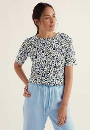 Vrouw met donker haar die gouden oorringen draagt, een korte mouwen bloemen top en lichtblauwe broek met hoge taille, tegen een effen achtergrond.