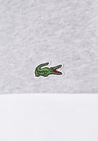 Logo de crocodile vert brodé avec un détail rouge sur fond de tissu gris et blanc, probablement sur un vêtement.