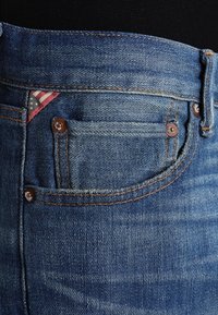 Los jeans de mezclilla azul presentan un diseño tradicional con estilo de cinco bolsillos, remaches de cobre y una tela estampada que muestra rayas rojas y blancas.