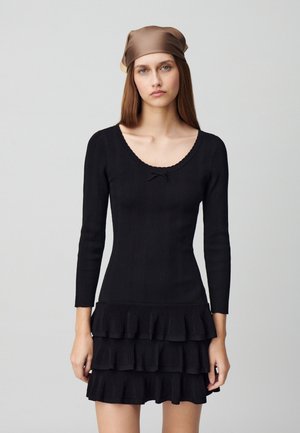 Robe noire en tricot côtelé avec des manches longues et un col à volants, comportant un ourlet à volants en trois couches. Texture lisse et silhouette ajustée.