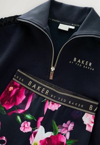 Námornícky zip-up top s kvetinovým potlačovým panelom a ružovými a zelenými akcentmi. Obsahuje čierny golier a zlaté písmená značky na látke.