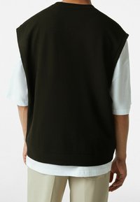 Černý top bez rukávů s texturovaným povrchem a vysokým výstřihem, vrstvený přes bílou košili, v kombinaci se světle béžovými kalhotami.