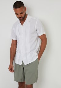 Camisa blanca de manga corta y botones, hecha de una tela ligera, que presenta rayas verticales, con un corte relajado, combinada con pantalones cortos de color verde oliva.