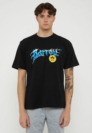 Giovane uomo con capelli ricci scuri che indossa una maglietta nera con scritta blu "Barrow" e una faccina gialla con denti aguzzi, pantaloni grigio chiaro e tatuaggi sulle braccia.