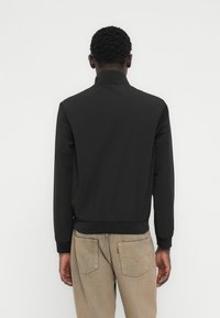 Άνδρας φορώντας μαύρο μπουφάν με ψηλό γιακά και μπεζ τζιν, στέκεται με την πλάτη στην κάμερα μπροστά σε απλό λευκό φόντο.