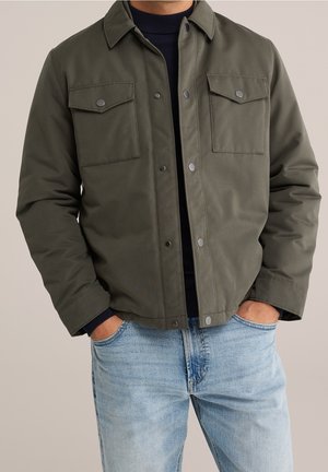 Mies pukeutuneena oliivinvihreään takkiin, jossa on painonapit ja rintataskut, mustan villapaidan päällä, siniset farkut, kädet taskuissa.