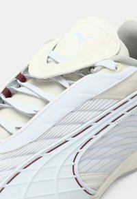 Biele a béžové tenisky Puma s zakrivenými dizajnovými detailmi, bielymi šnúrkami a viditeľným logom Puma na jazyku.