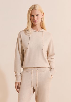 Femme blonde portant un sweat à capuche en maille beige et un pantalon à cordon assorti, debout avec une main dans la poche devant un fond neutre.