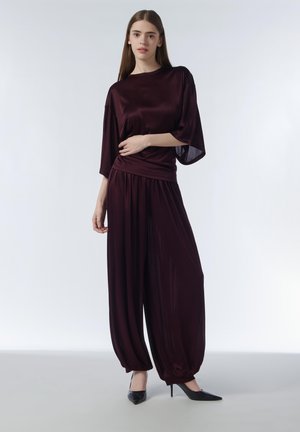 Young woman wearing a dark purple loose satin top and wide pants, standing with one arm across her waist, wearing black high heels.