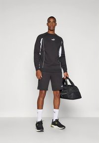 Homem vestindo sweatshirt e calções Puma pretos e brancos, meias brancas Puma, ténis pretos, segurando uma bolsa desportiva preta.