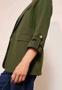 Olivově zelené sako s upraveným střihem, se štíhlým klopou, zapínáním na jeden knoflík a ohrnutými rukávy s akcenty knoflíků.