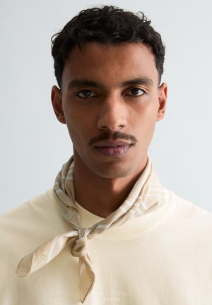 Beżowa koszulka z subtelnym logo, zestawiona z jasnym, pasiastym szalikiem zawiązanym wokół szyi. Krótkie, ciemne, kręcone włosy.