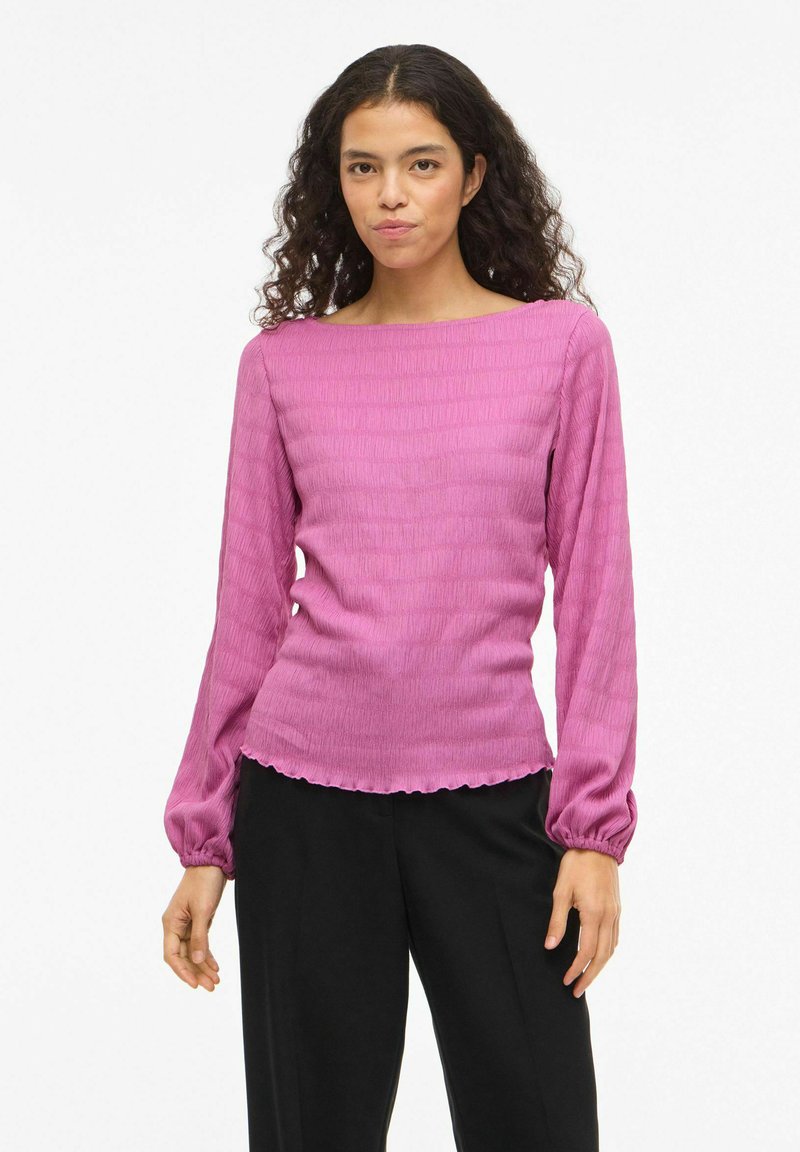 Langermet, fuksia topp med teksturert design og bølgete kant. Ermene er puffete ved håndleddene, noe som skaper en tettsittende silhuett.