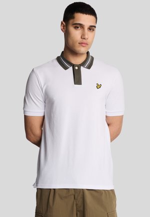Vit polo skjorta med en grårandig krage och en liten gul logotyp på bröstet. Korta ärmar och ett mjukt, strukturerat tyg.