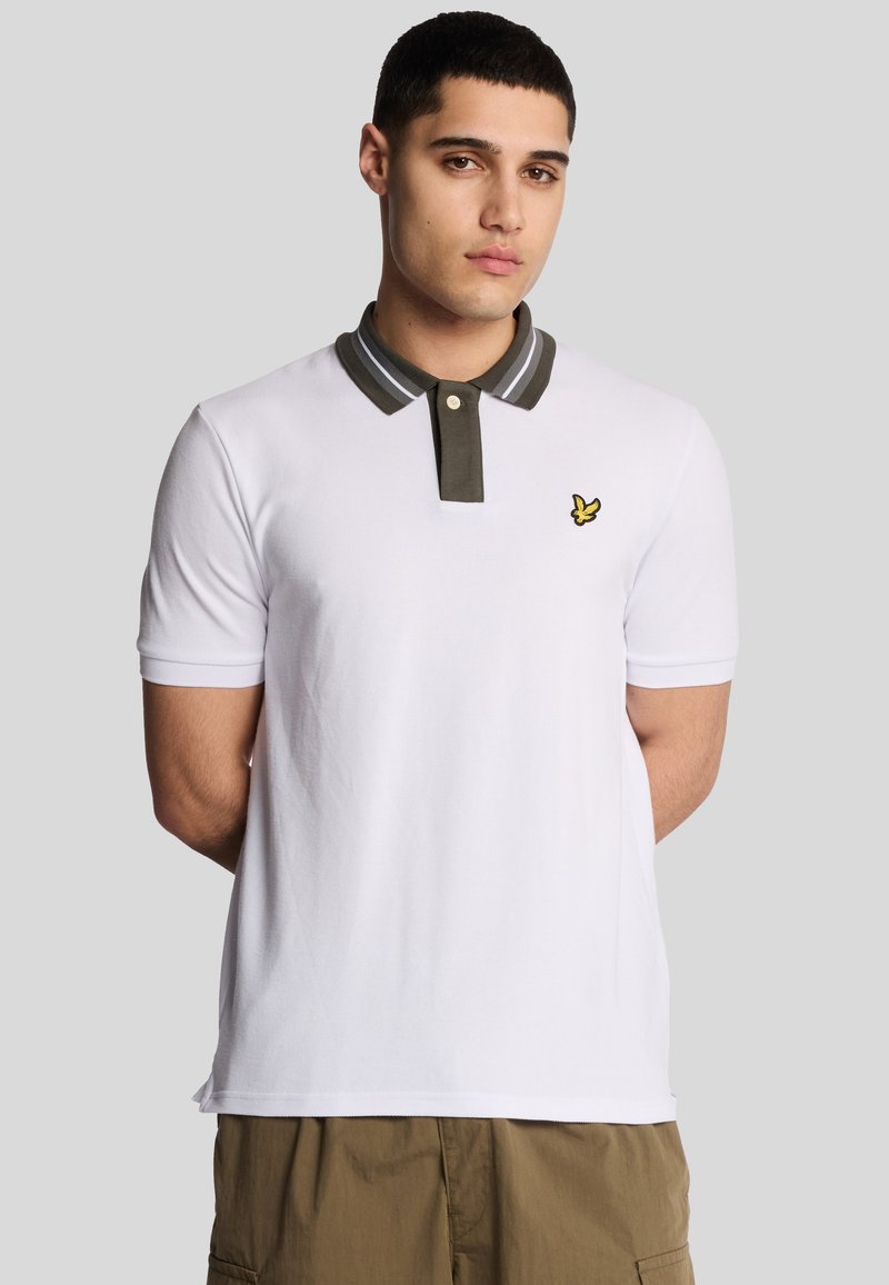 Hvid polo skjorte med en gråstribet krave og et lille gult logo på brystet. Korte ærmer og et blødt, tekstureret stof.