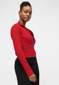 Junge Frau mit geflochtenem Haar trägt ein rotes langärmeliges Oberteil mit Reißverschluss und schwarze Hose und posiert vor einem einfarbigen, hellen Hintergrund.