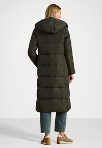 Garšs, olīvu zaļš pūkains mētelis ar kapuci, ar quiltētu dizainu un taisnu siluetu, saskaņots ar gaiši ziliem saīsinātiem džinsiem un bēšiem sneakeriem.