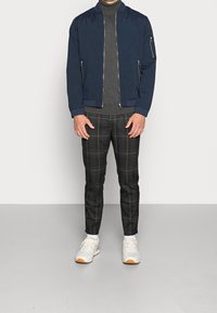 Granatowa kurtka bombowiec, szary sweter z golfem, spodnie w kratę w ciemnych odcieniach oraz białe trampki z pomarańczowymi akcentami. Metalowe zamki.