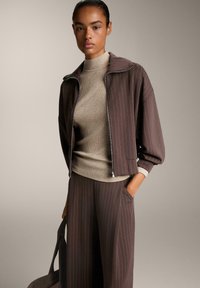 Kvinde i brun stribet jakke og bukser med en beige ribbet turtle neck, bærende en mørkebrun taske, mod en enkel neutral baggrund.