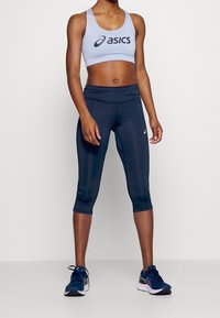 Sostenitore sportivo di colore azzurro chiaro con logo ASICS nero, abbinato a leggings alla caviglia blu scuro attillati e scarpe da corsa nere con un accento rosa.