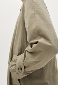 Beige, leichte Jacke mit lockerer Passform, ausgestattet mit einem knöpfbaren Manschette und strukturiertem Stoff. Taschen sind entlang der Seitennähte sichtbar.