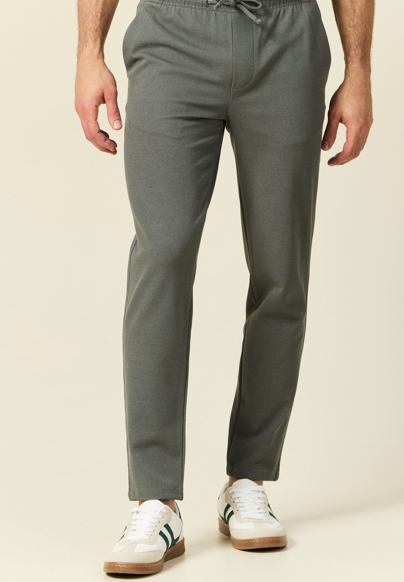 Grijze, taps toelopende joggers gemaakt van soepel materiaal met een elastische tailleband en koord, gecombineerd met witte sneakers met groene accenten.