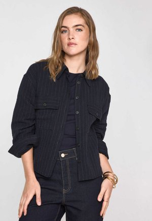Giacca camicia a righe navy con chiusura a bottoni e due tasche sul petto, abbinata a un top navy e pantaloni in denim scuro, completata da accessori dorati.