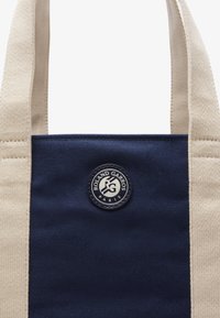 Sac fourre-tout en toile marine avec des poignées beiges ; présente un logo circulaire avec le texte "Roland Garros" et "Paris" au centre.