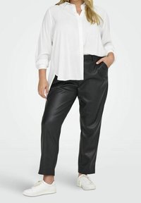 Donna che indossa una camicia bianca a maniche lunghe e oversize, pantaloni di pelle neri, sneakers bianche, con una mano in tasca, su uno sfondo semplice chiaro.