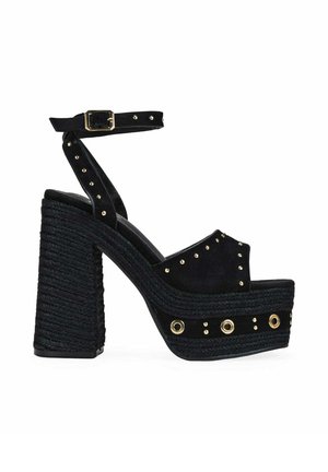 Sandalia de plataforma negra con tacón alto, textura tejida, correa en el tobillo, tachuelas doradas y detalles con ojales en el tacón y la suela gruesos.