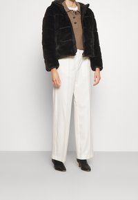 Svart fuskpälsjacka i cropped modell med huva, dragkedjestängning och horisontellt kviltad textur, kombinerad med lösa vita byxor och svarta ankelskor.