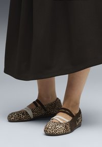 Sko med leopardtryk og beige samt brune accenter, med flad sål og krydsede stropper, placeret mod en sort nederdel.