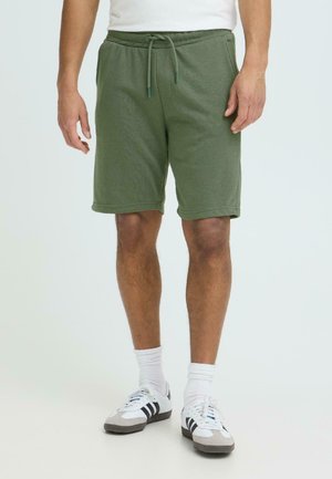 Mann trägt olivgrüne Kordelzug-Shorts, weiße Socken und weiße Adidas-Sneaker mit schwarzen Streifen, steht vor einem einfarbigen Hintergrund.
