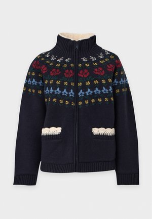 Une femme porte un cardigan bleu marine orné de bandes florales colorées, un haut noir, une jupe plissée couleur moutarde et des bottes noires, sur un fond de studio blanc.
