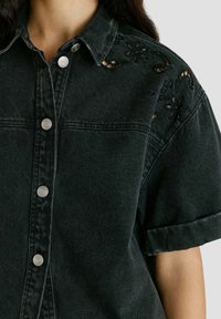 Chemise en denim noir à manches courtes avec boutons-pression argentés et broderie florale ajourée sur l'épaule portée par une personne.