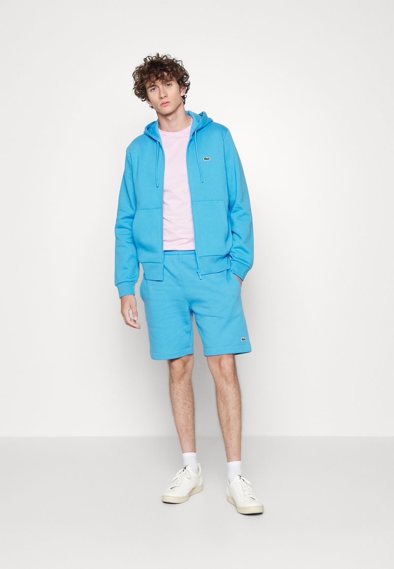 Niebieska bawełniana bluza z kapturem z przednimi kieszeniami, w zestawie z niebieskimi szortami i różową koszulką. Wyraźne logo Lacoste na lewej piersi i szortach.