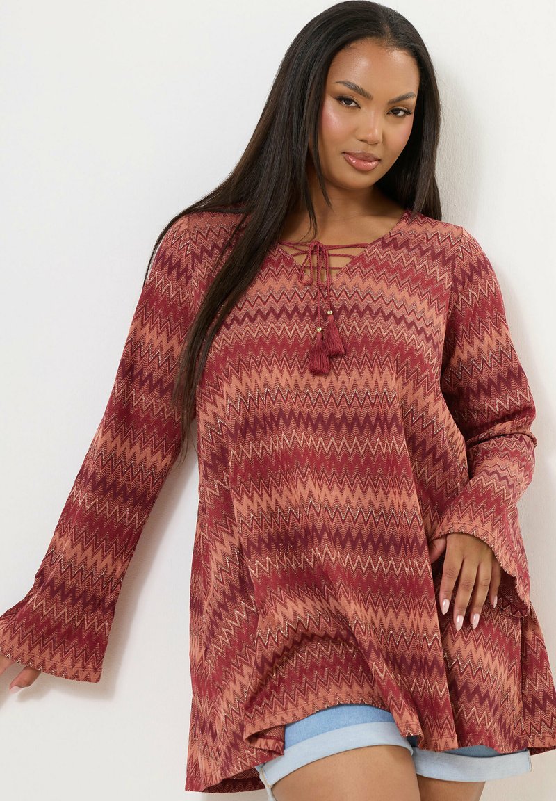 Tunika a maniche lunghe in motivo zigzag rosso e beige, con scollatura a V dotata di dettaglio con lacci e frange. Tessuto morbido con vestibilità rilassata.