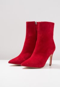 Czerwone zamszowe botki za kostkę z ostrym noskiem, obcasem w kształcie szpilki i pionowym zapięciem, charakteryzujące się miękką teksturą i smukłą sylwetką.