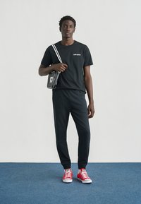 T-shirt en coton noir avec le logo "Converse", associé à un pantalon noir. Baskets rouges avec semelles blanches. Petit sac noir avec des accents blancs.