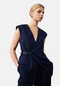 Marineblaues ärmelloses Wickeloberteil mit tiefem V-Ausschnitt, seitlichen Taschen und einem Gürtel mit silbernen Beschlägen für eine anpassbare Passform. Glatter, maßgeschneiderter Stoff.
