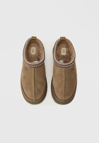 Brūni zamša čības ar mīkstu kažokādas odere, kontrastējoši šuvju akcenti ap kaklu un aizmugurē esoša cilpa vieglai uzvilkšanai. UGG zīmols redzams.